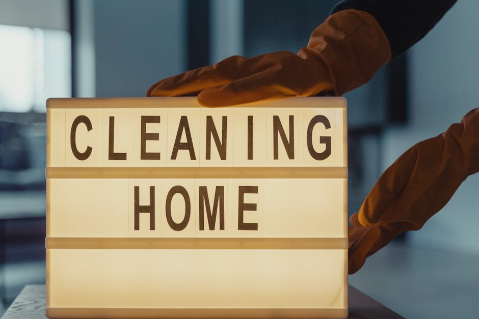 A person wearing brown cleaning gloves is placing a lit-up sign with black letters on a white background that reads 'CLEANING HOME'. The sign is positioned on a white surface in an indoor setting with